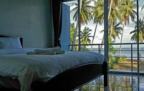 Een slaapkamer met een groot bed en uitzicht op palmbomen en de zee door een raam.