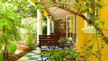 A covered outdoor seating area with a checkered floor, surrounded by greenery and a yellow wall.