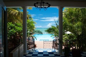 Blick von einer überdachten Terrasse auf einen Pool mit Palmen und blauem Himmel.
