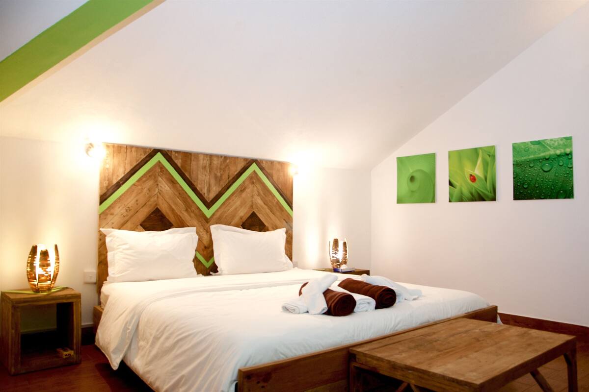 A neatly made bed with white linens and brown towels, a wooden headboard with a chevron pattern, and a small wooden table beside the bed.