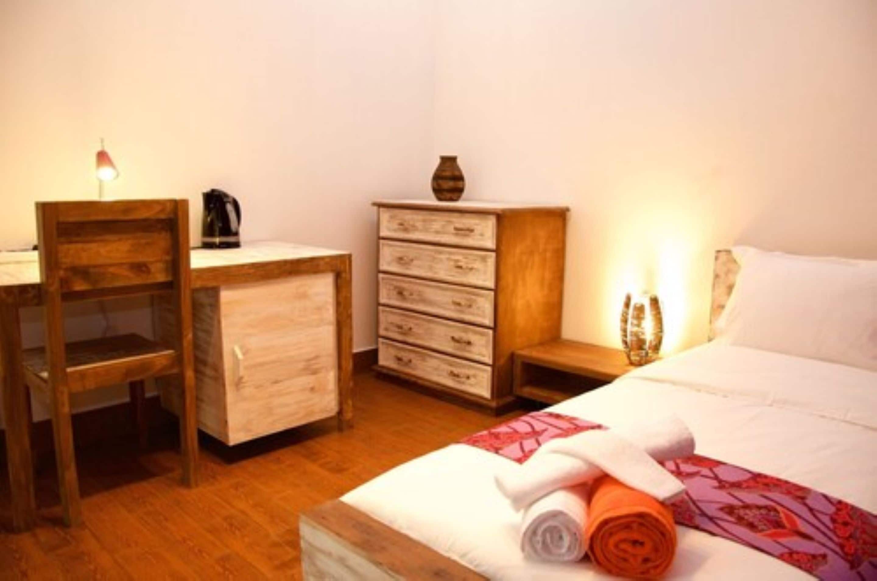 A bedroom with a wooden desk, a dresser, a bed with rolled towels, and a nightstand with a lamp.