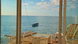 Vista desde una habitación con un ventanal grande que da a un mar tranquilo, un pequeño bote en el agua y una terraza de madera con dos sillas.