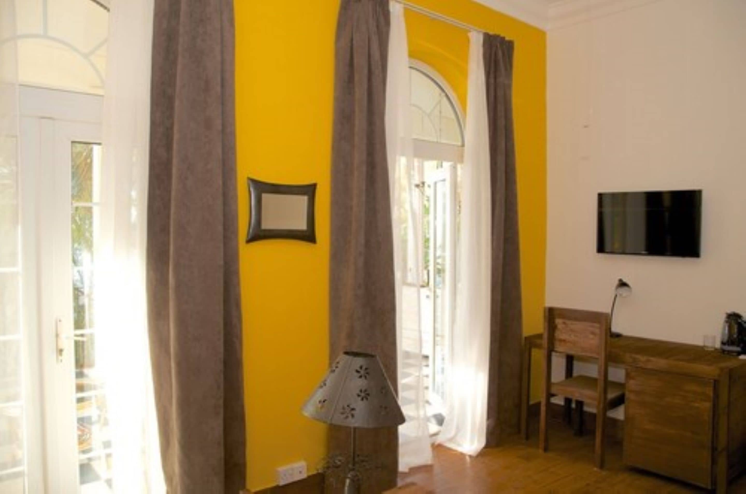 A room with yellow walls, a wooden desk, a chair, a lamp, and a TV mounted on the wall.