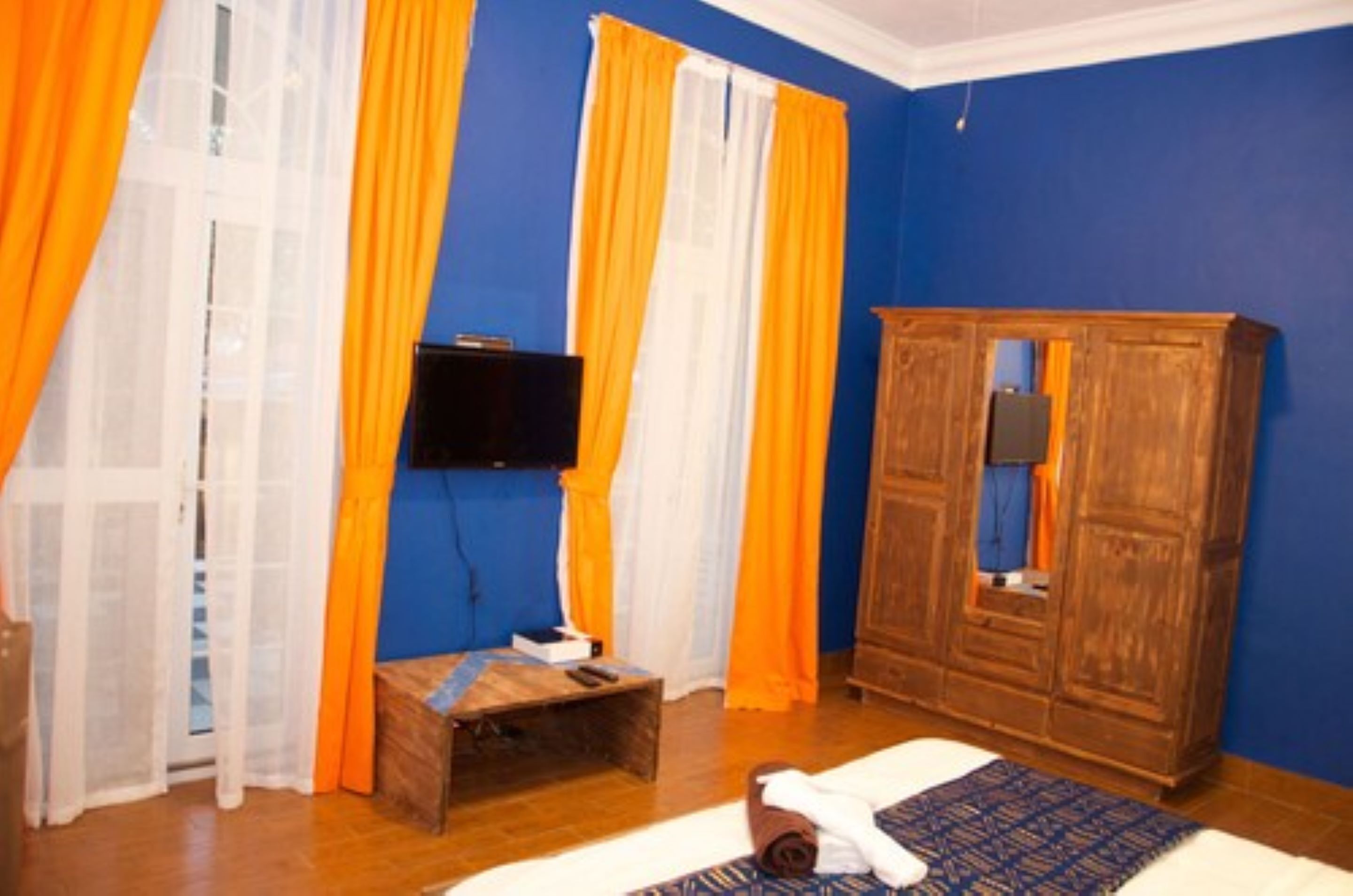 A room with blue walls, a wooden wardrobe, a television mounted on the wall, and a small wooden bench.