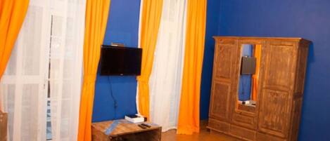 A room with blue walls, a wooden wardrobe, a television mounted on the wall, and a small wooden bench.