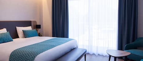 Una habitación de hotel moderna con una cama grande, un banco, una mesita y vistas a la ciudad a través de cortinas translúcidas.