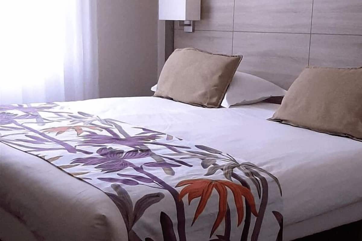 Habitación de hotel con cama, dos almohadas, cabecero y ventana con cortinas.