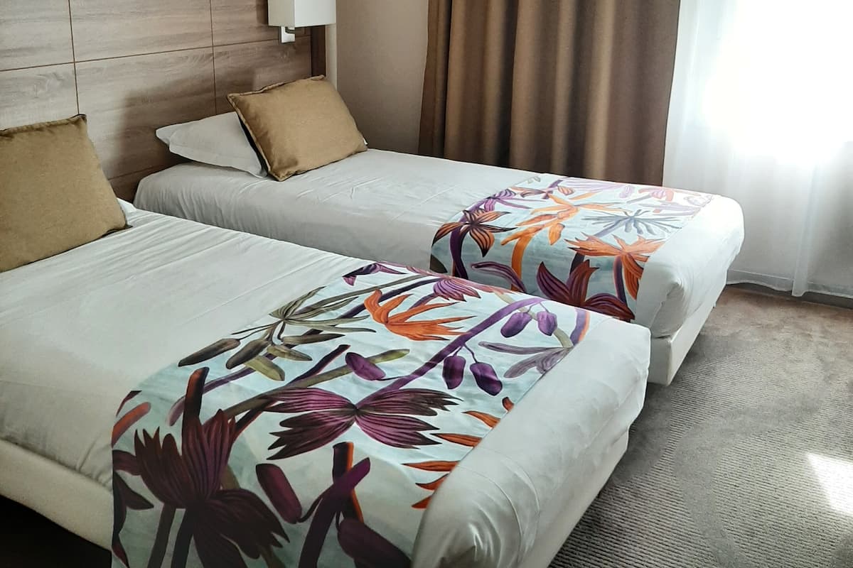 Habitación de hotel con dos camas, cabecera de madera, mesita de noche, lámpara y ventana con cortinas.