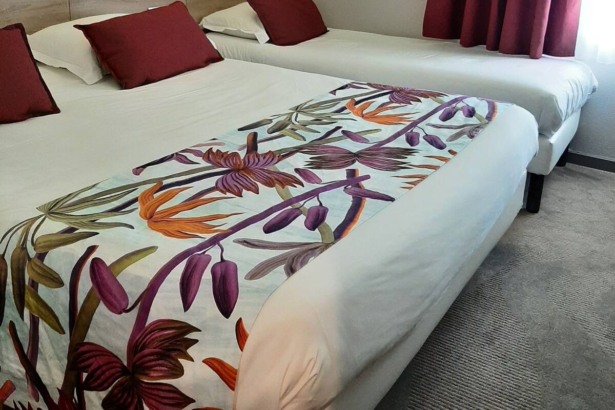 Habitación de hotel con dos camas, almohadas rojas y blancas, colcha estampada con motivos florales, cabecero de madera y ventana con cortinas rojas.