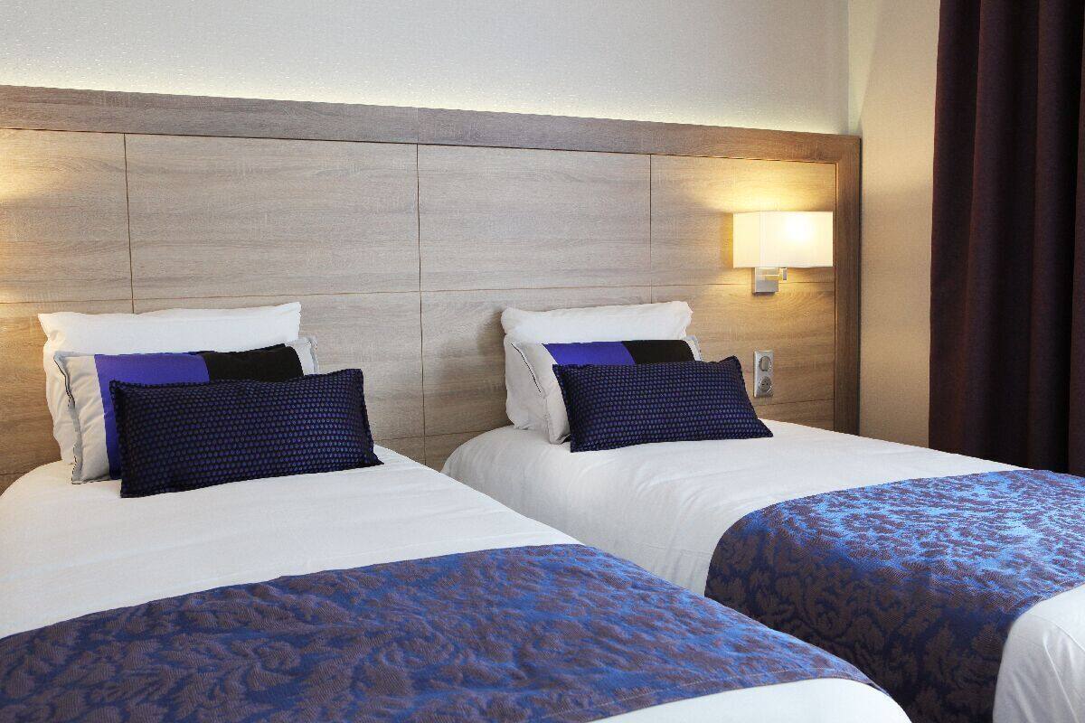Habitación de hotel con dos camas, cabecero de madera, lámpara de pared y ventana con cortinas.