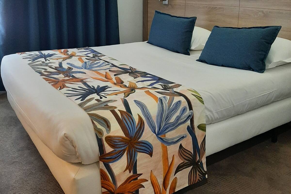 Una habitación de hotel con una cama, cabecero, lámpara de noche y ventana con cortinas.