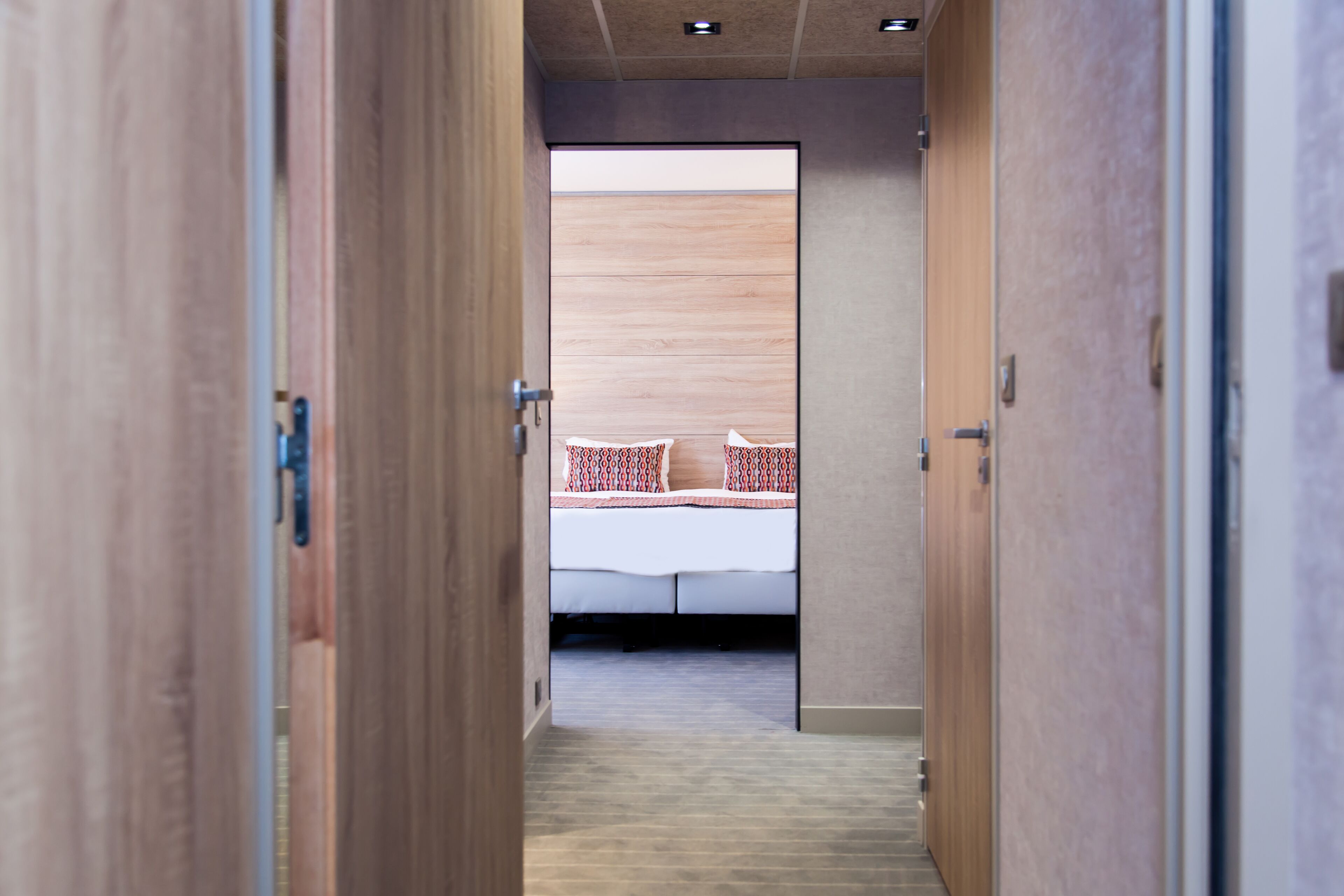 A hotel corridor leading to a room with a bed, wooden walls, and a carpeted floor.