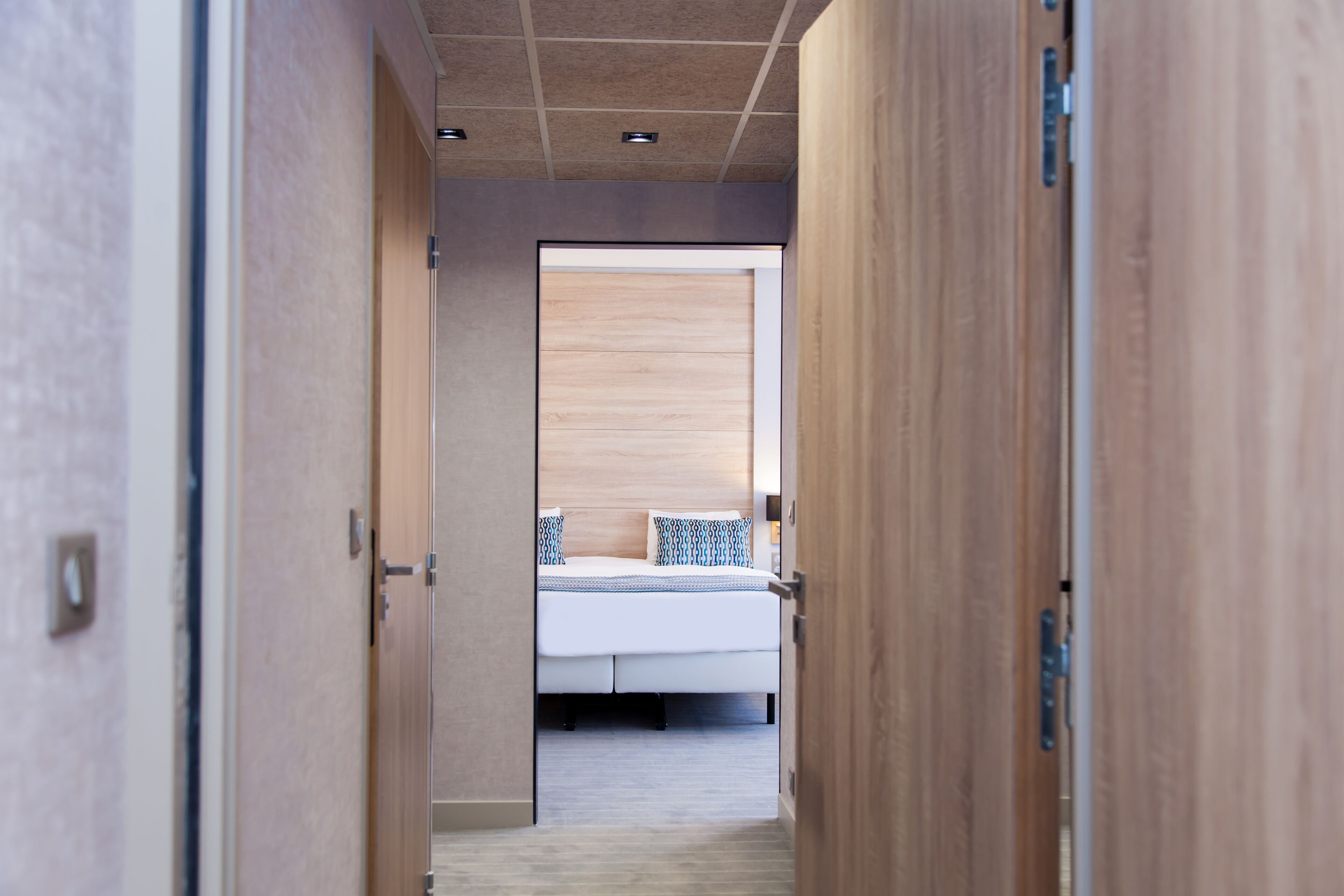 A narrow hallway leading to a bedroom with a bed, wooden doors, and a ceiling with recessed lighting.