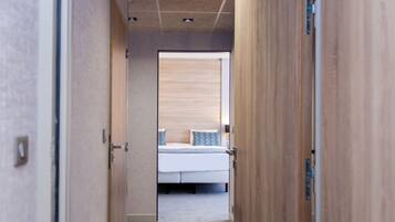 Ein schmaler Flur führt zu einem Schlafzimmer mit Bett, Holztüren und einer Decke mit eingelassenen Leuchten.