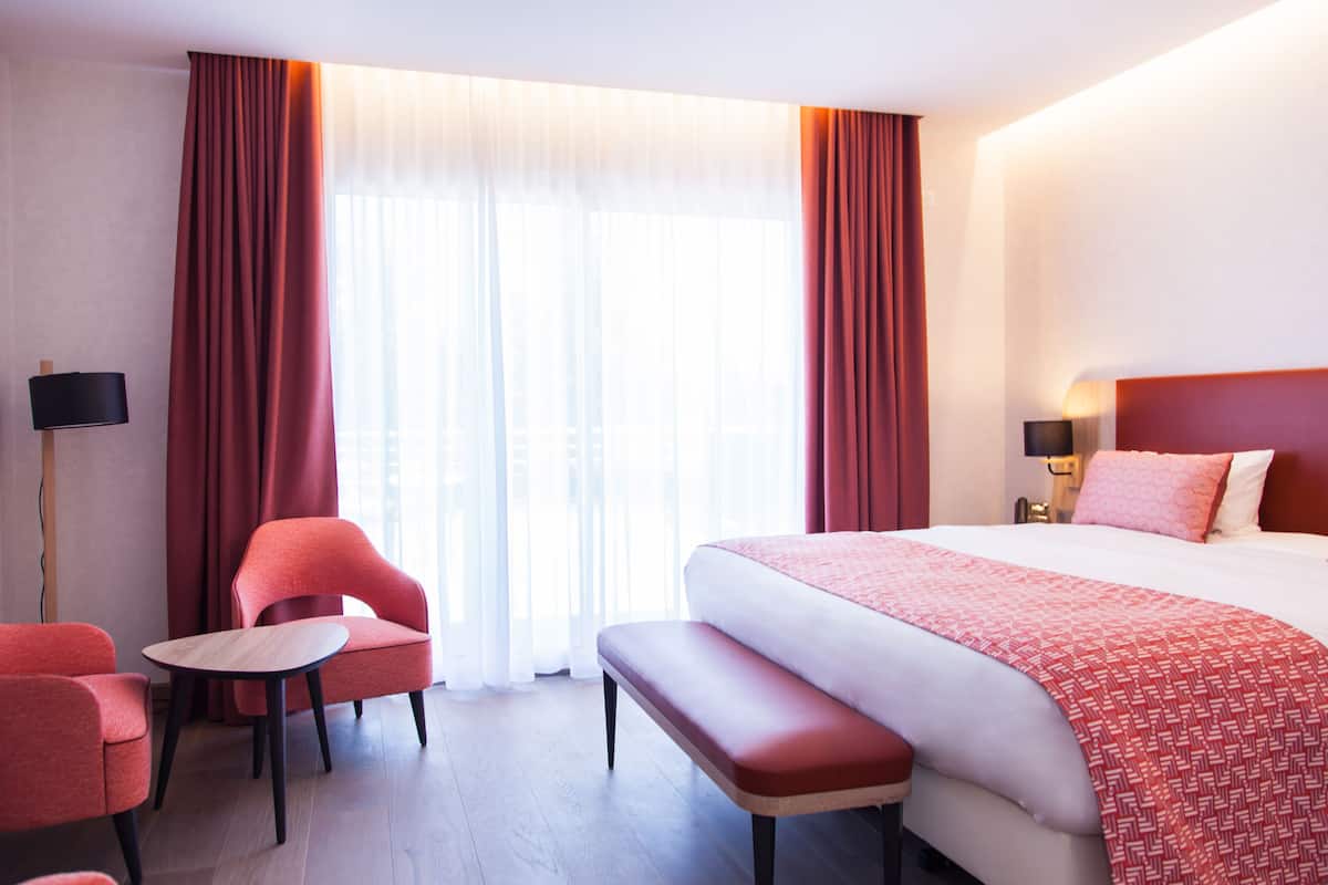 Una habitación de hotel con una cama grande, un banco, dos sillones, una mesa y una lámpara.