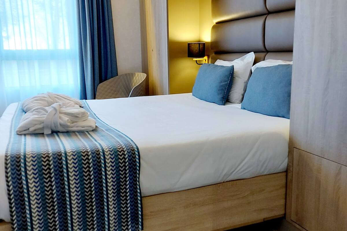 Habitación de hotel con una cama grande, una mesita de noche, una lámpara y una ventana con cortinas.