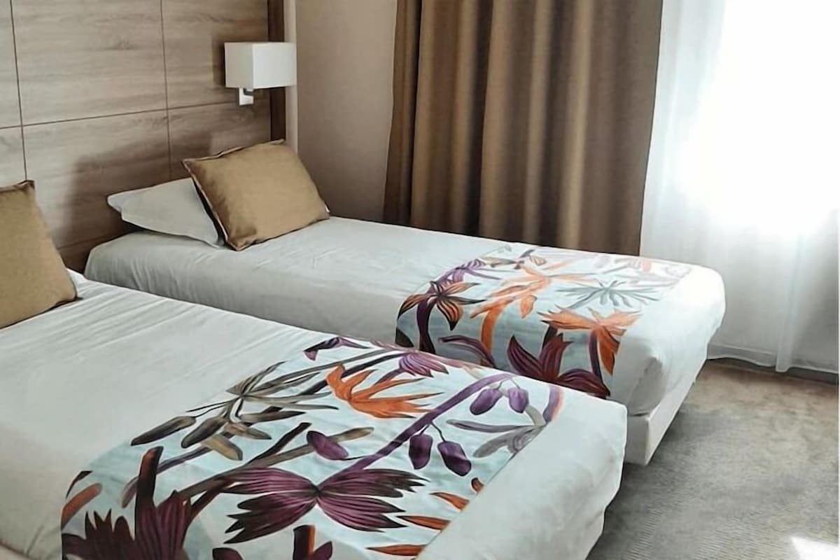 A hotel room with two beds, a headboard, a bedside lamp, and a window with curtains.