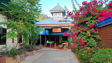 A courtyard with a building featuring a blue roof and a decorative white facade. There are wooden tables and chairs outside, and a variety of plants and flowers around.