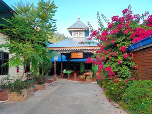 A courtyard with a building featuring a blue roof and a decorative white facade. There are wooden tables and chairs outside, and a variety of plants and flowers around. - Boondee House (Mae Hong Son)
