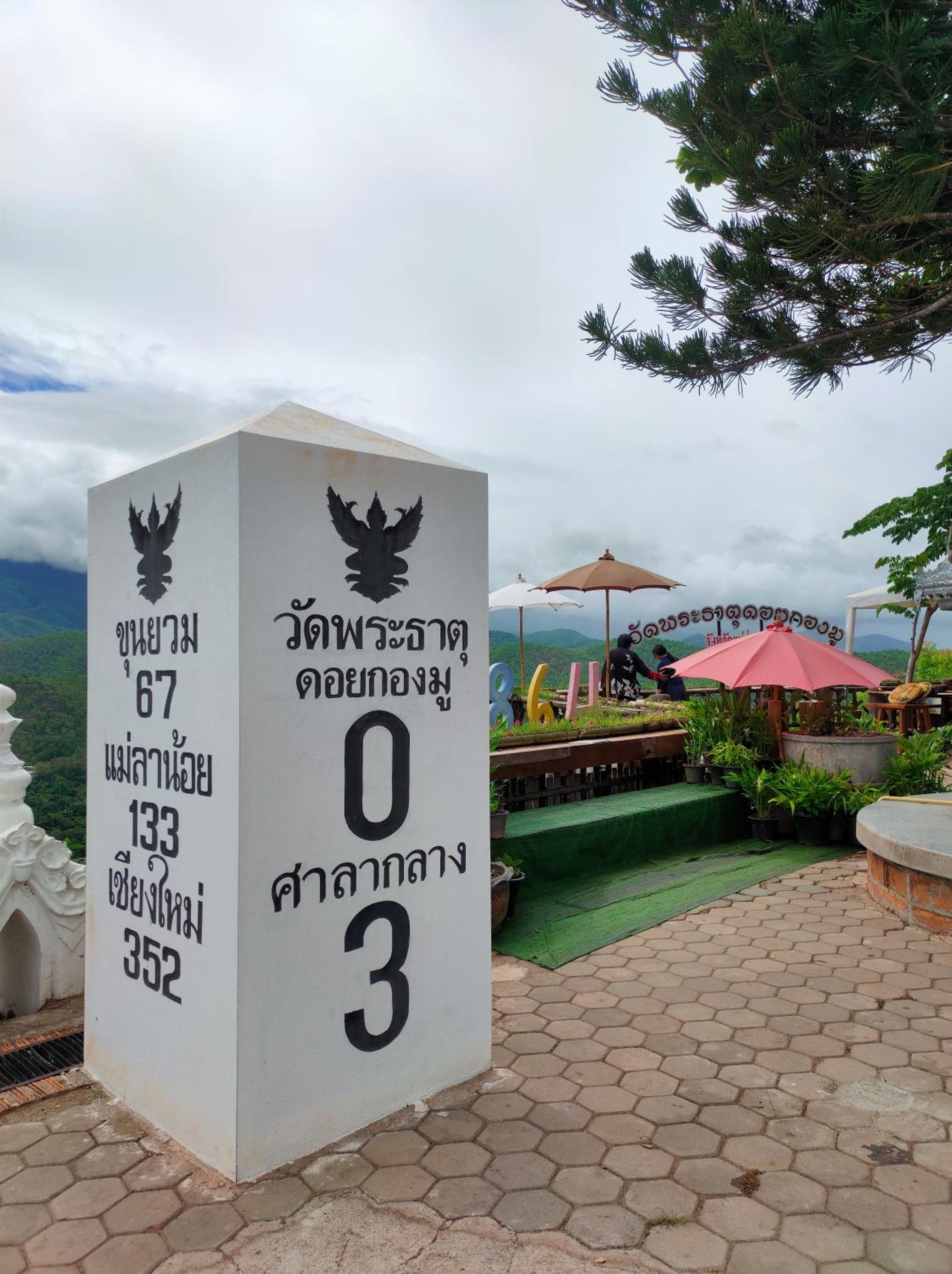 A white pillar with Thai text and numbers, surrounded by a paved area with umbrellas and potted plants.
