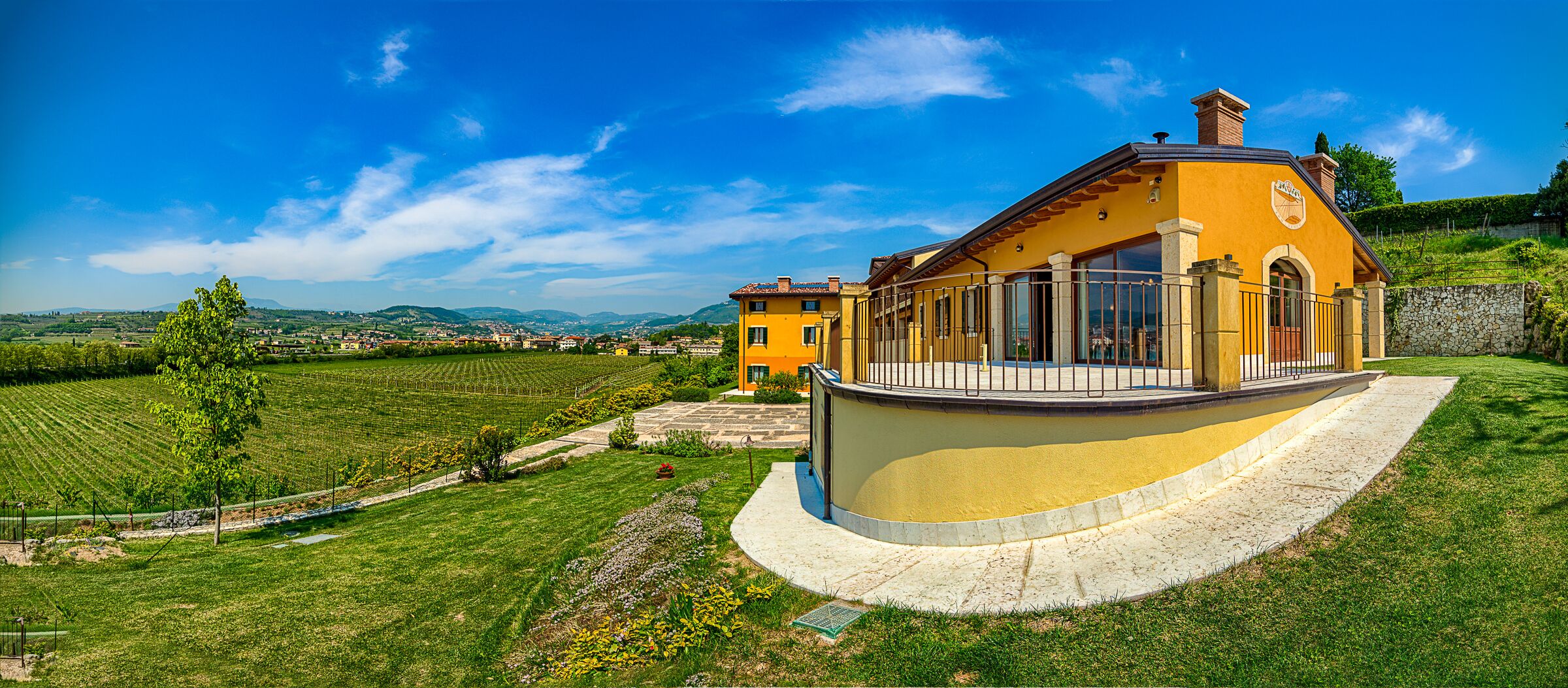 A yellow building with a curved balcony, surrounded by a well-maintained garden and vineyards.