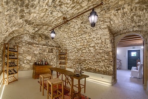 A stone-walled room with a wooden table and chairs, a wine rack, and a bookshelf.