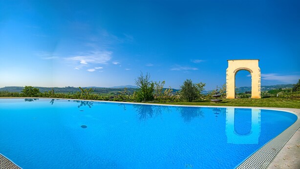 Piscina azul cristalina con una fuente, rodeada de follaje y una gran estructura en forma de arco.