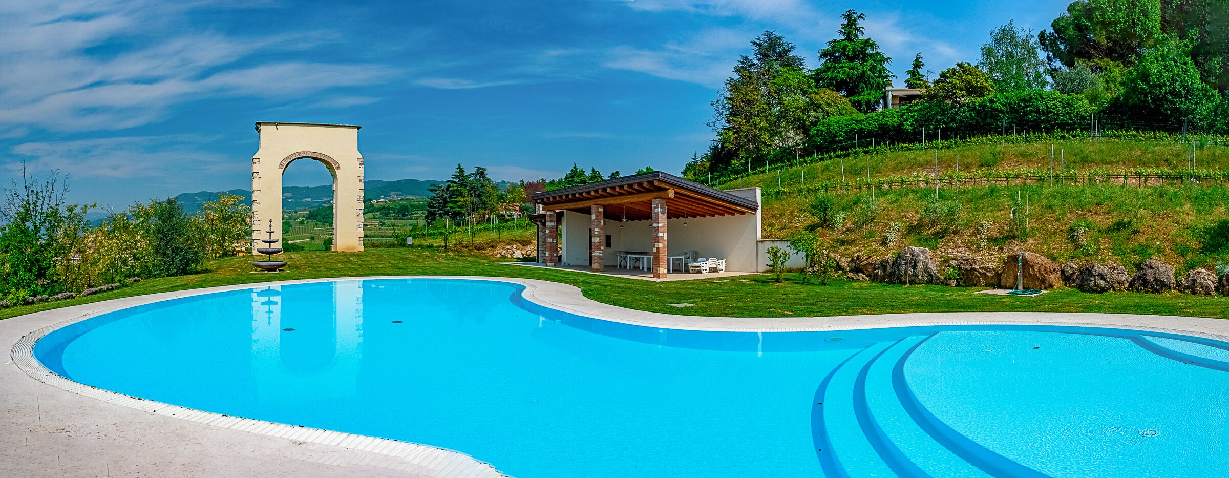 Ein Schwimmbecken mit einem Steingewölbe, einer überdachten Terrasse und einer sanften, grasbewachsenen Böschung.