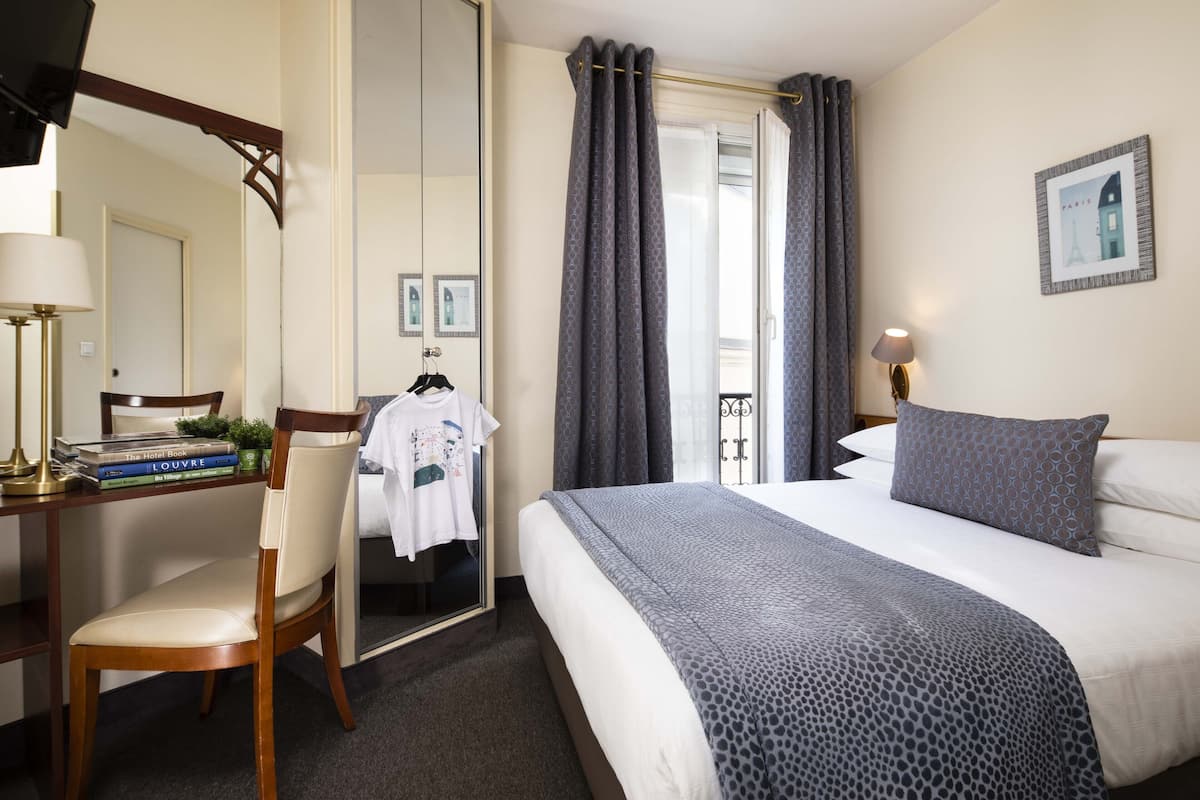 A hotel room with a bed, a desk with a chair, a mirror, a wardrobe, and a window with curtains.