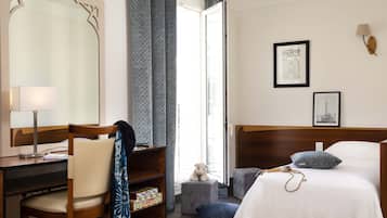Habitación de hotel con un escritorio de madera, una silla y una cama con edredón blanco. Hay una ventana con cortinas, una lámpara de pared y dos cuadros en la pared.