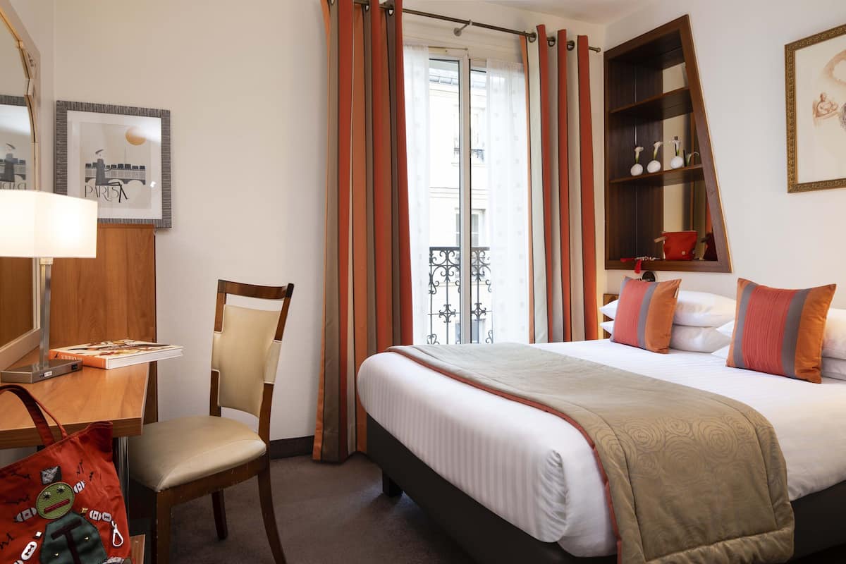 A hotel room with a bed, a chair, a desk, a lamp, a shelf with decorative items, and a window with curtains.