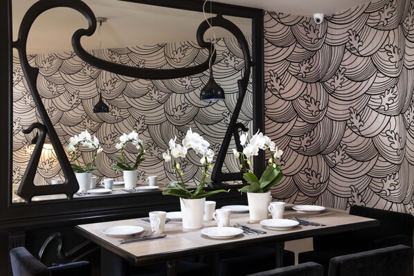 Un comedor con una mesa dispuesta para cuatro personas, platos y tazas blancos, y un jarrón con orquídeas blancas. Un espejo grande refleja la habitación, y el papel tapiz de la pared tiene un diseño de olas.