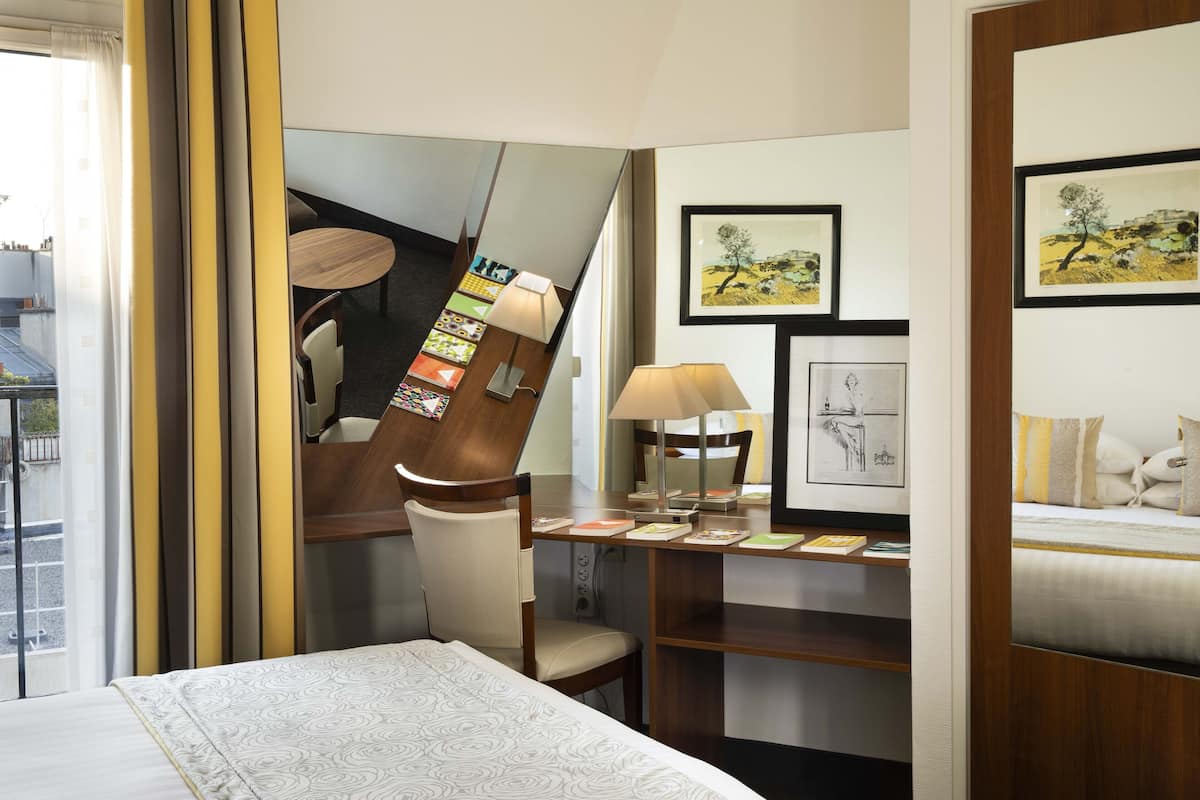 A hotel room with a bed, a desk with a chair, a bookshelf, and artwork on the walls.