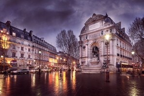 A historic building with classical architecture, a central monument, and a wet street reflecting lights. - Grand Hotel des Balcons (Paris)