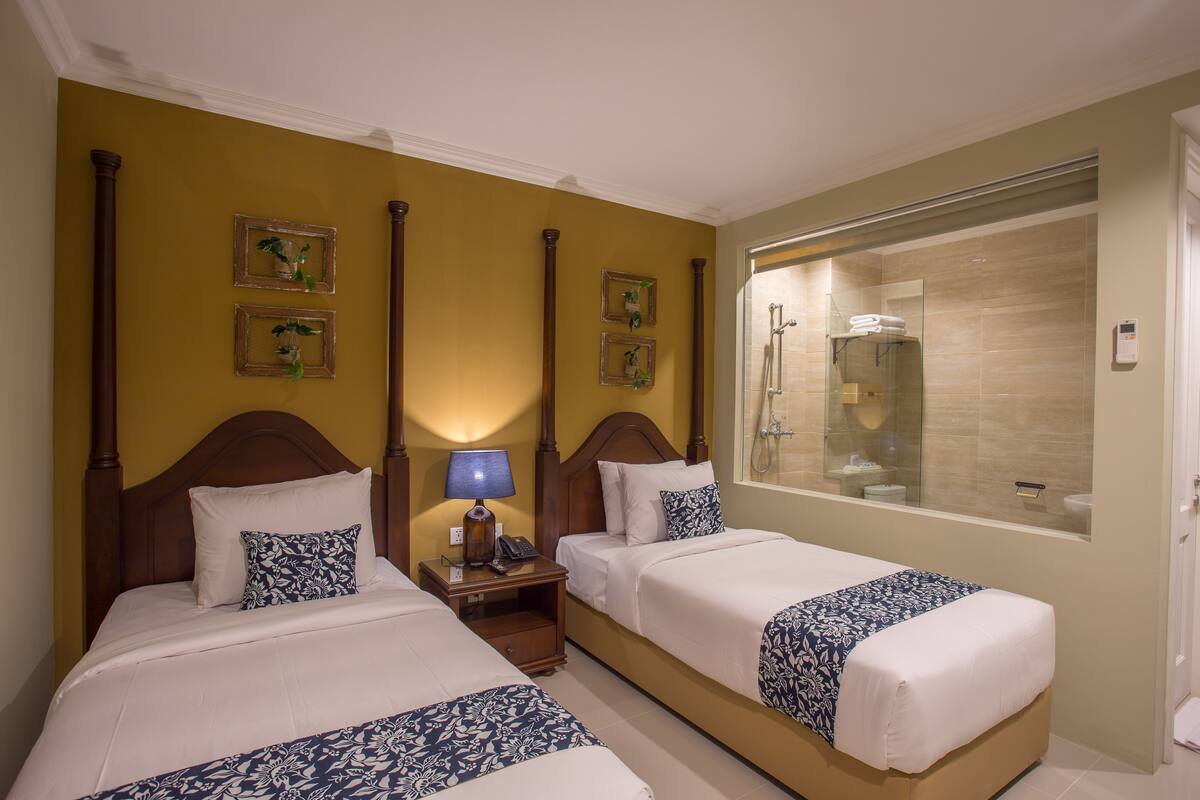 A hotel room with two beds, a nightstand with a lamp, a bathroom with a glass shower enclosure, and framed pictures on the wall.