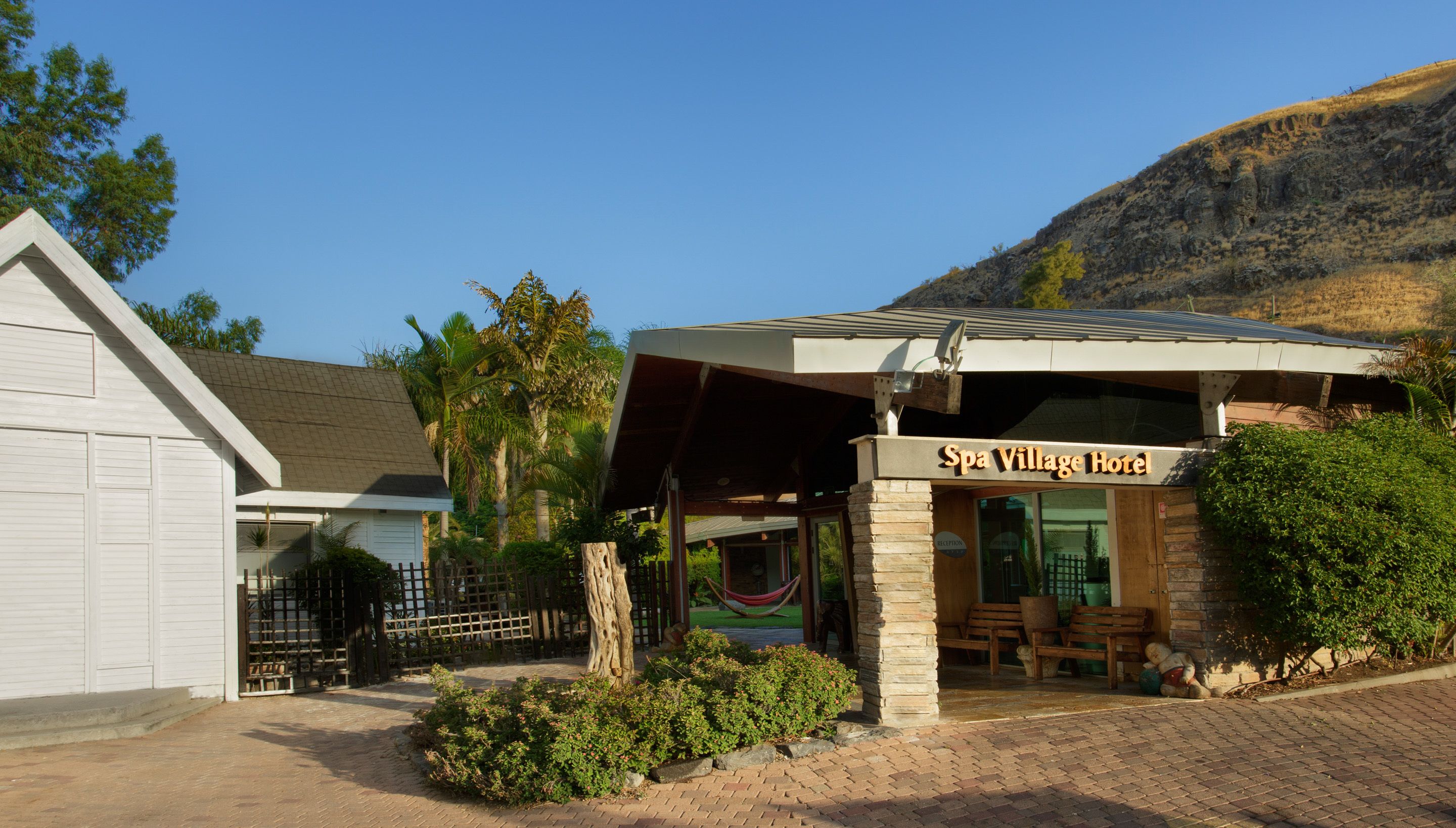 A building labeled "Spa Village Hotel" with a covered entrance, outdoor seating, and a mountain backdrop.