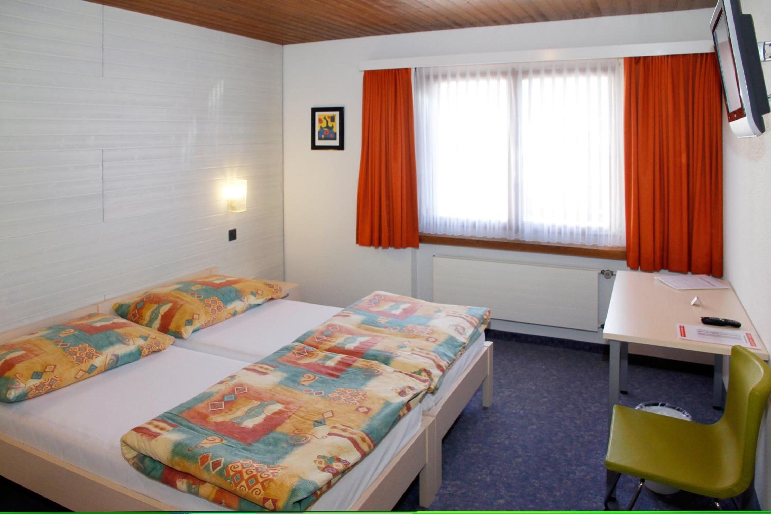 A room with two beds, a desk, a chair, a window with orange curtains, and a wall-mounted TV.