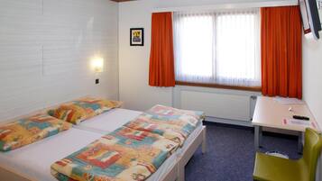 A room with two beds, a desk, a chair, a window with orange curtains, and a wall-mounted TV.