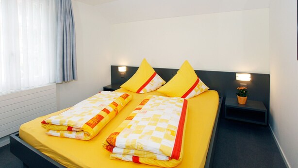 A double bed with yellow bedding and two pillows, a bedside table with a lamp, and a window with sheer curtains. - Hotel Krone (Attinghausen)