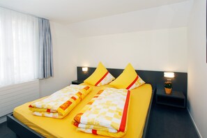 Ein Doppelbett mit gelber Bettwäsche und zwei Kissen, ein Nachttisch mit Lampe sowie ein Fenster mit transparenten Vorhängen.