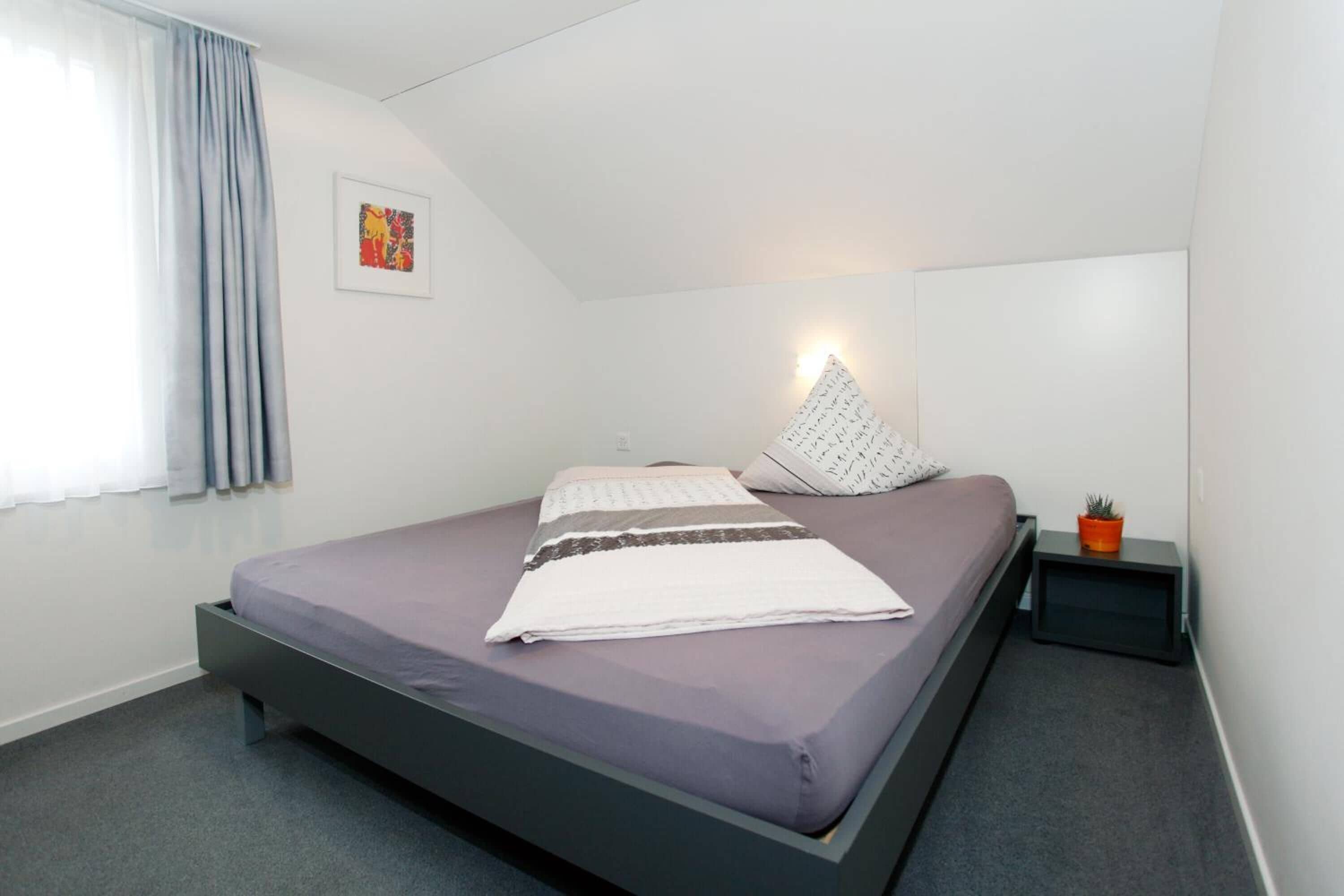Ein modernes Schlafzimmer mit einem Bett, einer Nachttischlampe, einem kleinen Tisch mit einer Pflanze und einem Bild an der Wand.