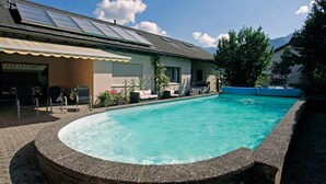 A swimming pool with a stone edge, a covered patio area with outdoor furniture, and a building with a sloped roof and solar panels. - Hotel Krone (Attinghausen)