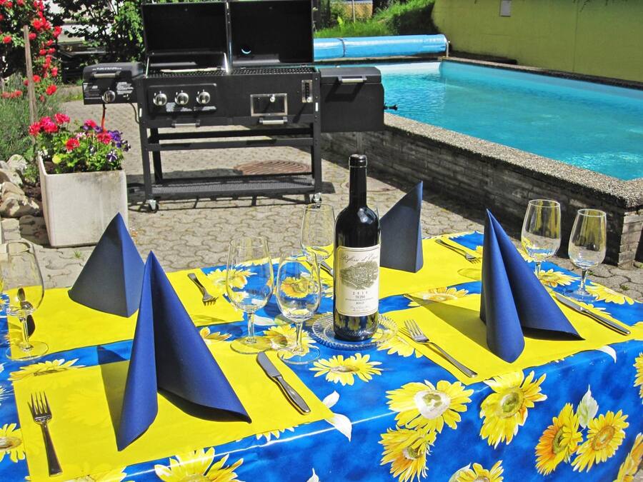Een terras met een eettafel gedekt voor twee personen, met een fles wijn, wijnglazen en een blauw-gele tafelkleed met bloemen.