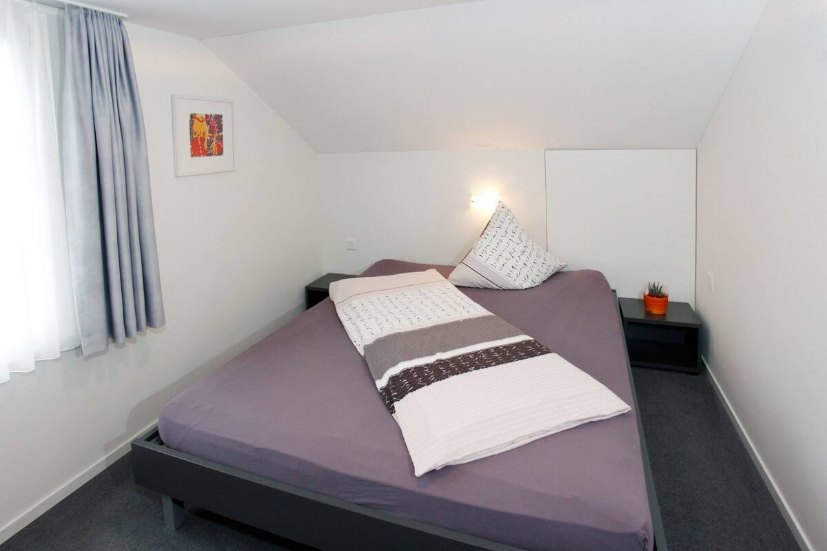 A single bed with a purple bedspread and multiple pillows, a bedside table with a plant, and a wall-mounted light.