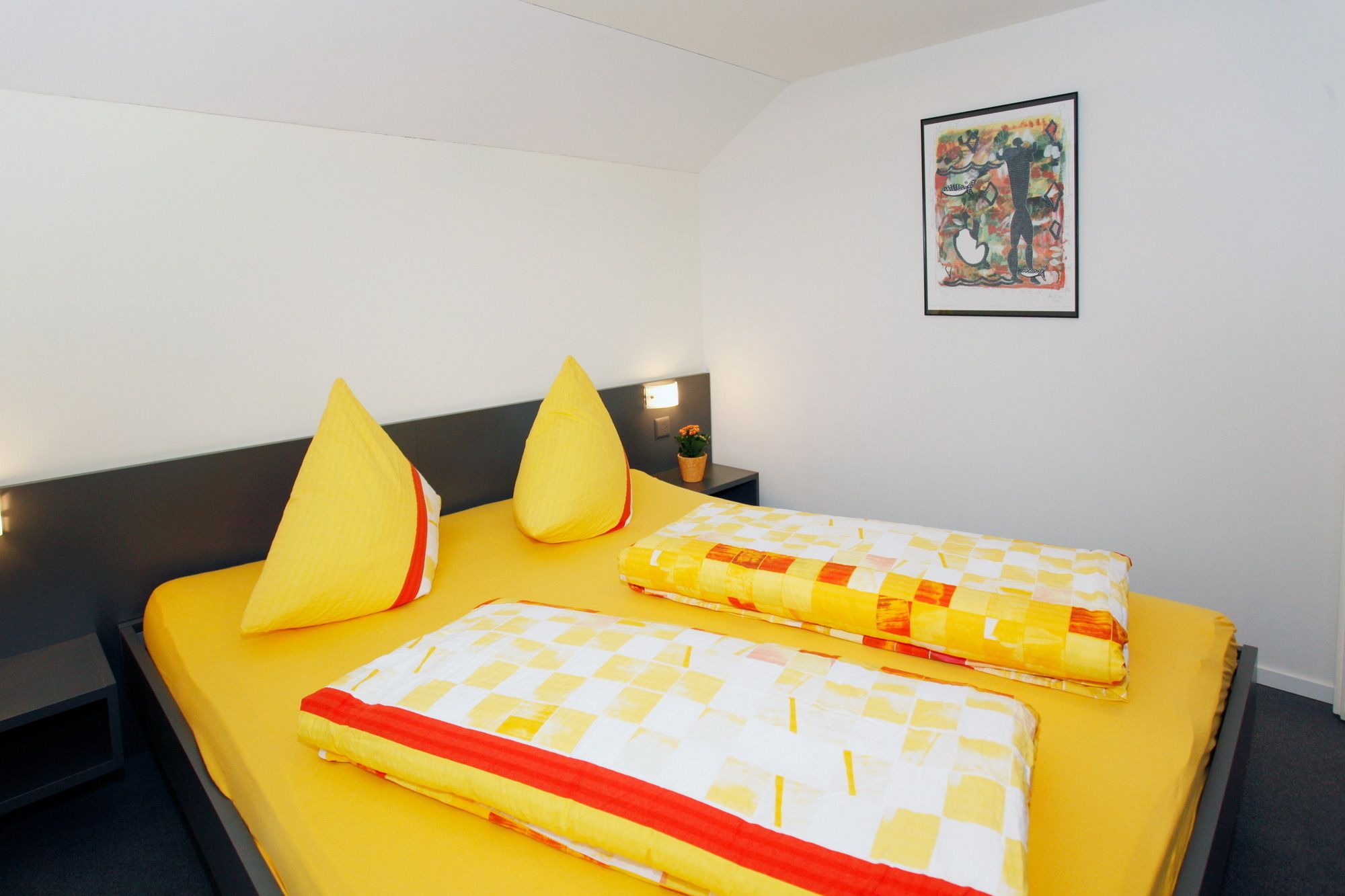 Une chambre d’hôtel avec un lit, deux oreillers, une table de chevet, une lampe fixée au mur et un tableau encadré.