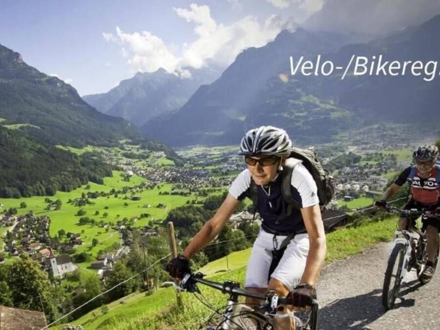 Dos ciclistas en una carretera de montaña con una vista panorámica de un valle y montañas al fondo.