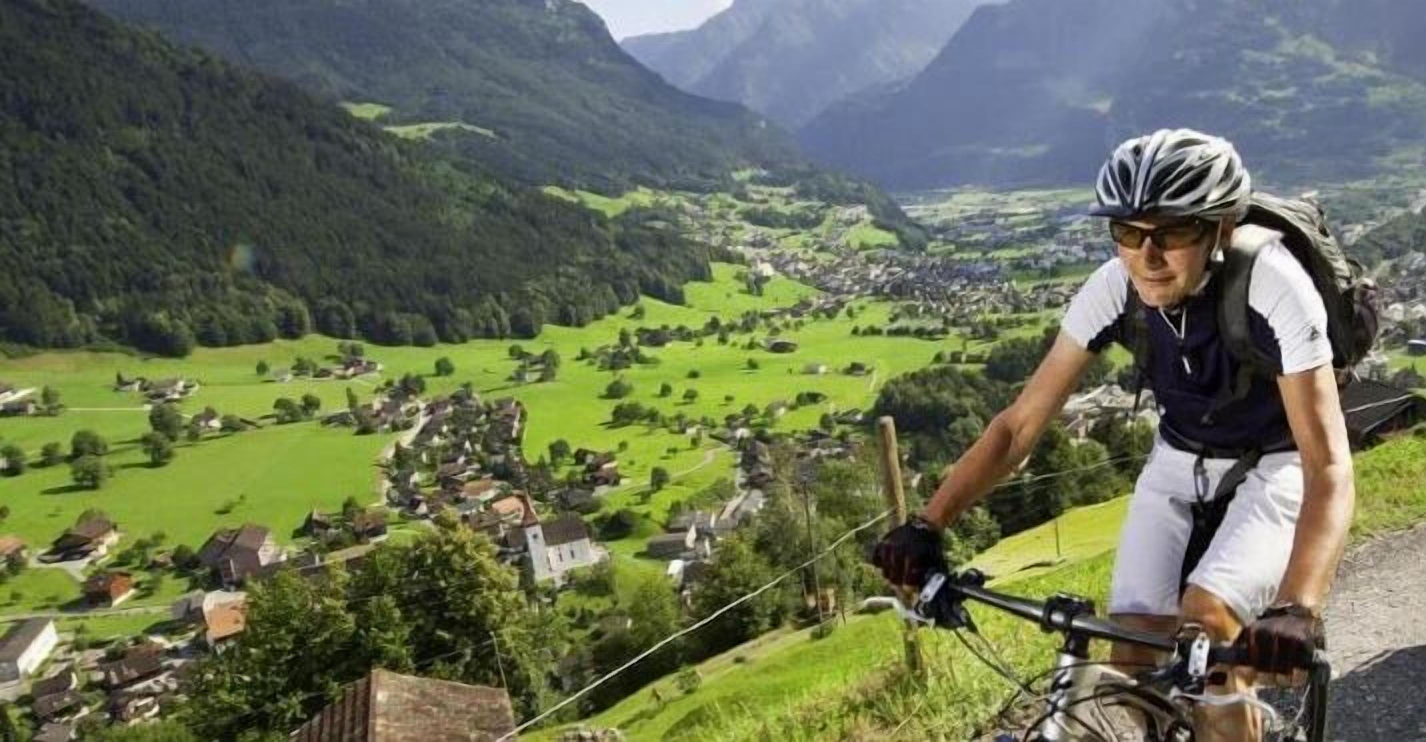 Un cycliste sur un sentier de montagne, avec vue sur une vallée et quelques maisons dispersées.