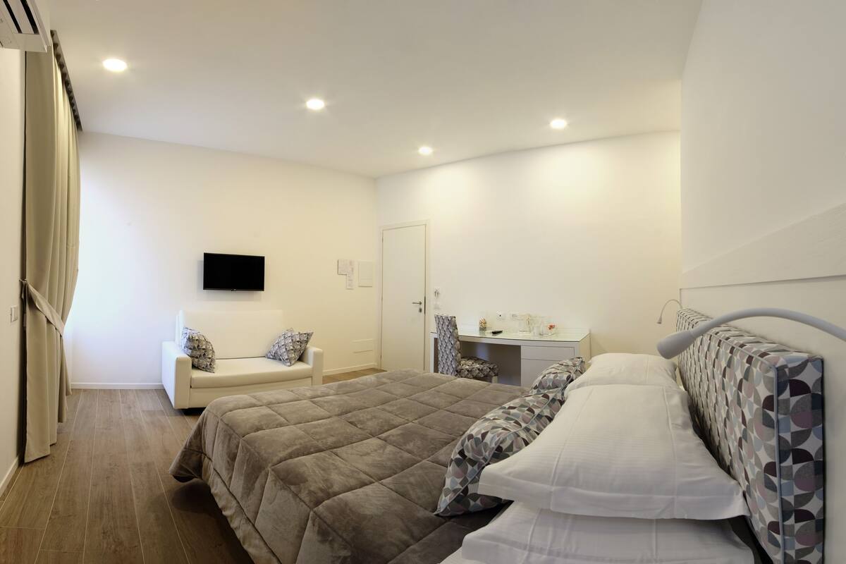 Un dormitorio con una cama grande, una mesita de noche, una silla, un escritorio y un televisor montado en la pared.