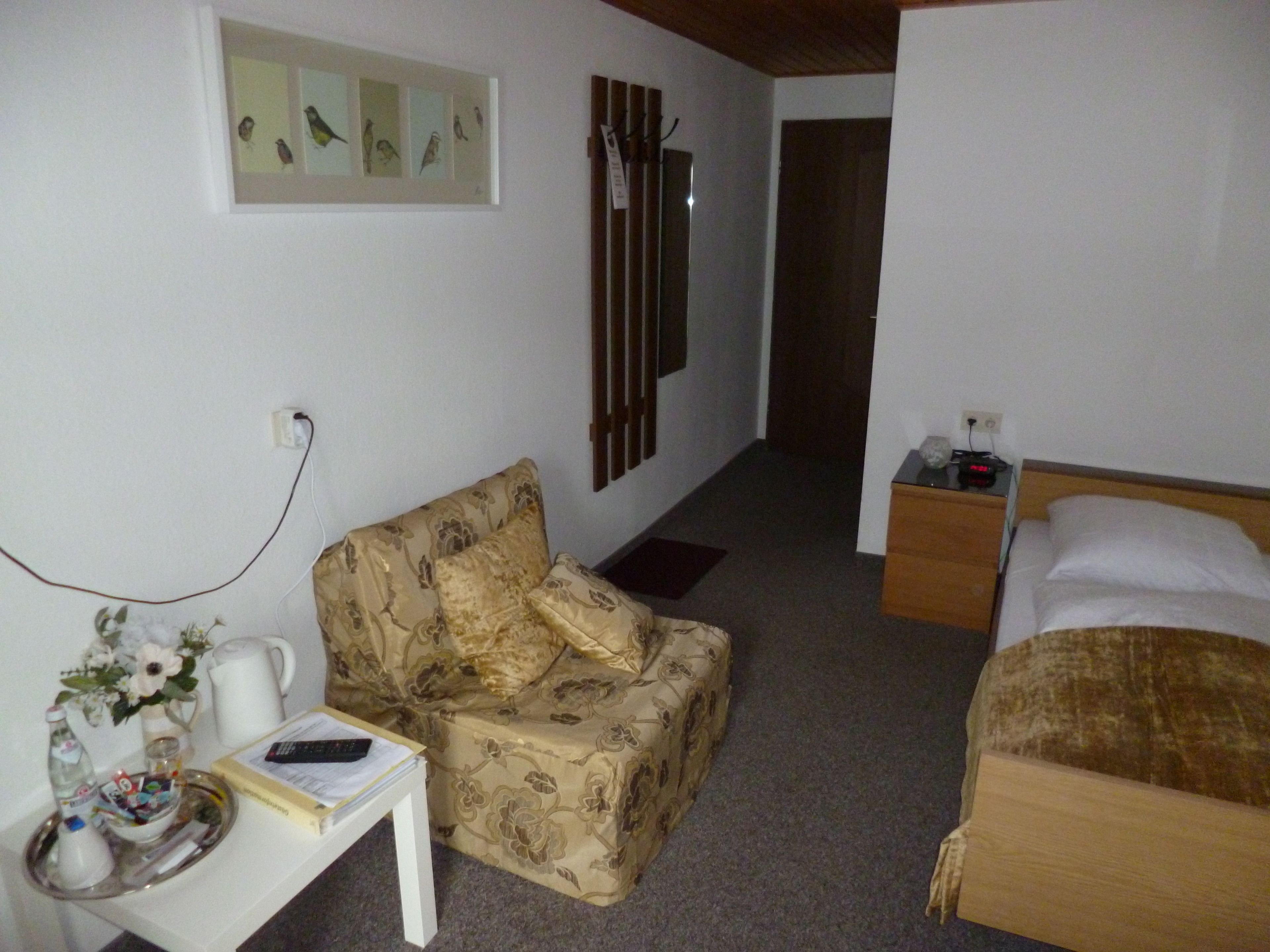 A room with a bed, a sofa, a small table with a remote control, and a wall-mounted shelf with pictures.