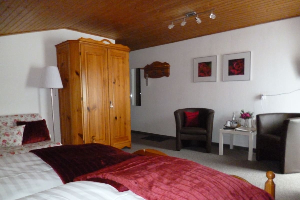 Ein Schlafzimmer mit einem großen Holzschrank, einem Bett mit roter Decke, einem Sessel, einem kleinen Tisch mit einer Vase und zwei gerahmten Bildern an der Wand.